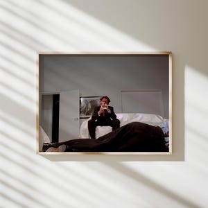 Könnte beinhalten: Ein gerahmter Druck, der einen Mann in einem schwarzen Anzug und roter Krawatte zeigt, der auf einem weißen Sofa sitzt. Die Szene umfasst einen teilweise sichtbaren Körper, der von einem schwarzen Laken bedeckt ist, und ein gerahmtes Kunstwerk an der Wand.