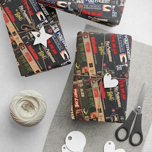 Könnte beinhalten: Mehrere Geschenkpakete mit Geschenkpapier, das mit VHS-Filmcovern bedruckt ist, darunter "The Exorcist" und "Re-Animator". Jedes Paket ist mit Bindfaden gebunden und hat ein stern- oder herzförmiges Etikett. Ein Knäuel Bindfaden und eine Schere sind ebenfalls vorhanden.