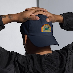 May include: A navy blue baseball cap featuring a semi-circular embroidered patch on the front. The patch displays a landscape graphic with a sun, hills, and the text "UCL ROSTOV" and "190284" in yellow, framed by orange and blue borders.