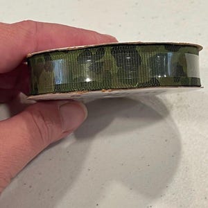 May include: A roll of camouflage ribbon. The ribbon is a dark green color with a repeating pattern of black and brown shapes. The ribbon is wrapped around a cardboard spool and is held in place with clear plastic.