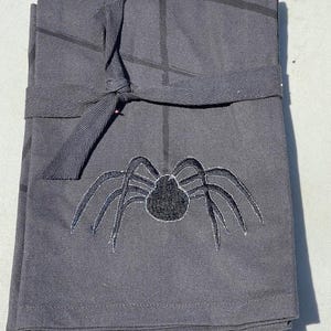 May include: A stack of gray cloth napkins tied with a matching ribbon. Each napkin features a black spider embroidered on the front. The napkins have a grid pattern and are suitable for Halloween.