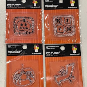 May include: Four clear stamps in plastic packaging. The stamps feature Halloween-themed designs: a pumpkin, buttons, a spider, and a bat. Each stamp is labeled "Snag 'em Stamps/Tampon Encreur" and "Spooky Town Collection". The packaging has an orange background.
