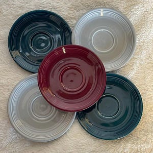 May include: Set of five ceramic saucers in various colors, including teal, gray, and burgundy. The saucers have a slightly raised rim and a smooth, glossy finish.