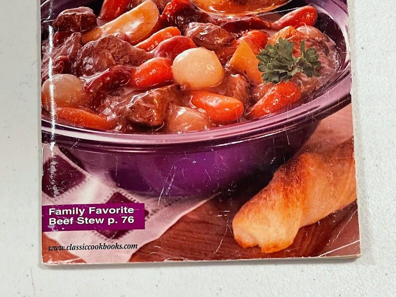 May include: A close-up of a cookbook page featuring a hearty beef stew. The stew is in a purple bowl and includes chunks of beef, carrots, and onions. A piece of bread is on the side. The text "Family Favorite Beef Stew p. 76" is visible.