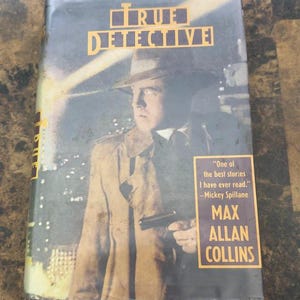 May include: A vintage paperback book titled "TRUE DETECTIVE" by Max Allan Collins. The cover features a man in a fedora and trench coat holding a gun. A quote from Mickey Spillane is also on the cover.