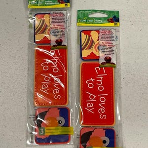 May include: Two packages of Sesame Street stickers. Each package contains a rectangular sticker that says "Elmo loves to play" in white text on a red background, and a square sticker with Elmo's face. The packages also include text that says "You can help, too!"