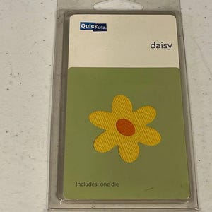 May include: A QuicKutz die set in a clear plastic package. The card inside features a yellow daisy with an orange center on a green background. The word "daisy" is printed above the flower. The text "Includes: one die" is printed at the bottom.