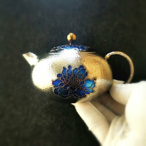 May include: Silver teapot with a hammered finish and a blue floral design. The teapot has a gold knob on the lid.