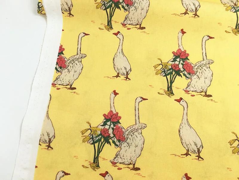 May include: Yellow fabric with a repeating pattern of white geese with red beaks and feet. Some geese are near red and pink floral arrangements. The fabric has a white border on the left side.