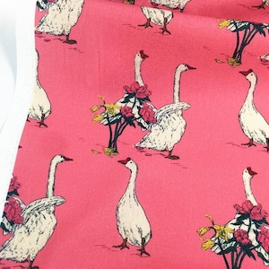 May include: Pink fabric with a repeating pattern of white geese with red beaks and feet. Some geese are adorned with floral arrangements of pink and yellow flowers. The fabric has a white edge on the left side.
