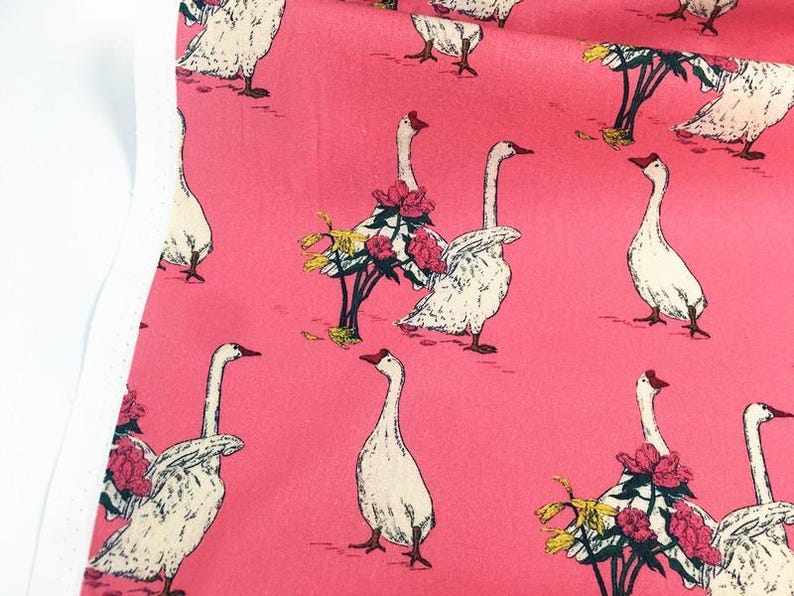 May include: Pink fabric with a repeating pattern of white geese with red beaks and feet. Some geese are adorned with floral arrangements of pink and yellow flowers. The fabric has a white edge on the left side.