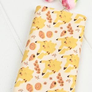 Könnte beinhalten: Gefalteter Stoff mit einem sich wiederholenden Muster aus gelben Zeichentrickfiguren, orangefarbenen Donuts und braunen Blattmotiven auf cremefarbenem Hintergrund. Der Stoff eignet sich für Bastel- oder Nähprojekte.