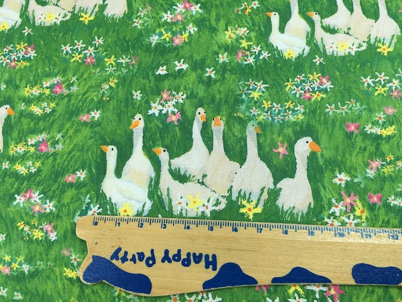 May include: Fabric with a green background featuring white geese with orange beaks and feet, amidst colorful flowers. A wooden ruler with the text "Happy Party" is at the bottom. The fabric is likely cotton or a similar material.
