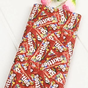 Op de afbeelding: Rode stof met een herhaald patroon van Skittles snoepverpakkingen. Het Skittles-logo en de snoepjes zijn zichtbaar. De stof heeft een heldere rode kleur.