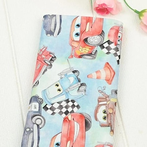 Könnte beinhalten: Hellblauer Stoff mit einem sich wiederholenden Muster aus Cartoon-Rennwagen, Leitkegeln und Zielflaggen. Die Autos sind rot, blau und braun, mit schwarz-weißen Zielflaggen. Geeignet für Kinderbasteln und Nähprojekte.