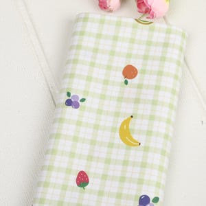 Könnte beinhalten: Ein gefalteter Stoffballen mit einem grün-weißen Vichy-Muster. Der Stoff ist mit Illustrationen von Früchten wie Banane, Orange, Erdbeere und Blaubeeren verziert. Der Stoff eignet sich für verschiedene Bastelprojekte.