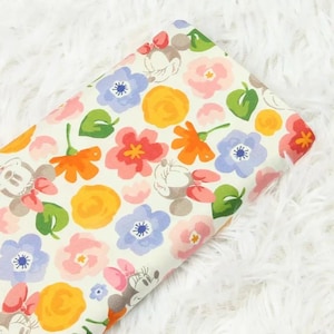 Peut inclure: Tissu à motifs floraux avec des illustrations de Minnie Mouse. Le motif comprend des fleurs orange, roses et bleues sur fond blanc. Le tissu est présenté sur une surface blanche et duveteuse.