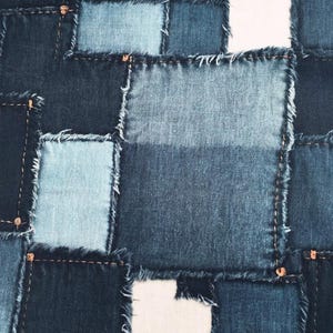 Może przedstawiać: Zbliżenie wzoru patchworku z kwadratami z niebieskiego dżinsu w różnych odcieniach. Kwadraty są zszyte widoczną nicią i mają postrzępione krawędzie, tworząc teksturowany, ręcznie robiony wygląd. Projekt obejmuje jaśniejsze i ciemniejsze odcienie dżinsu.