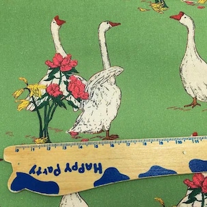 May include: Green fabric with a repeating pattern of white geese with red beaks and feet. Some geese are adorned with pink flowers and yellow blooms. A wooden ruler with the text "Happy Party" is in the foreground.
