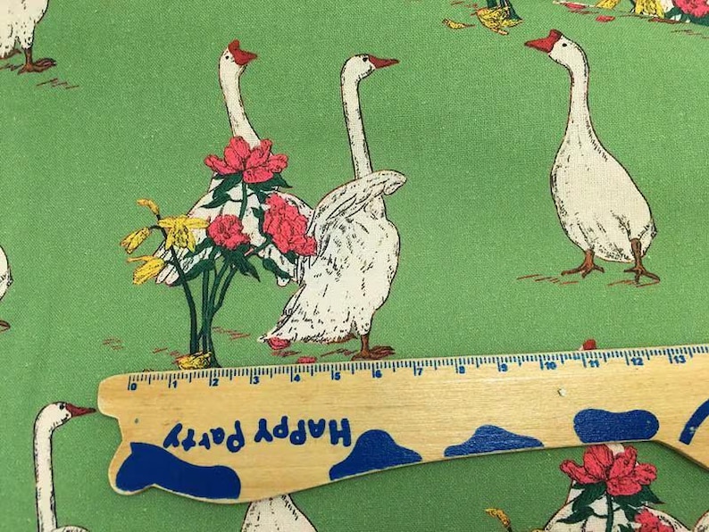 May include: Green fabric with a repeating pattern of white geese with red beaks and feet. Some geese are adorned with pink flowers and yellow blooms. A wooden ruler with the text "Happy Party" is in the foreground.