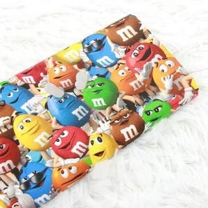 Peut inclure: Un tissu coloré avec un motif de bonbons M&M's de différentes couleurs et expressions. Les bonbons portent la lettre "m". Le fond est une surface blanche et duveteuse.