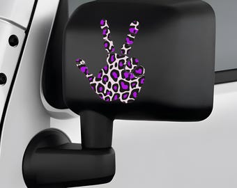 May include: A black side mirror with a peace sign decal. The peace sign is a hand gesture with a purple and white leopard print design. The mirror is attached to a white vehicle.