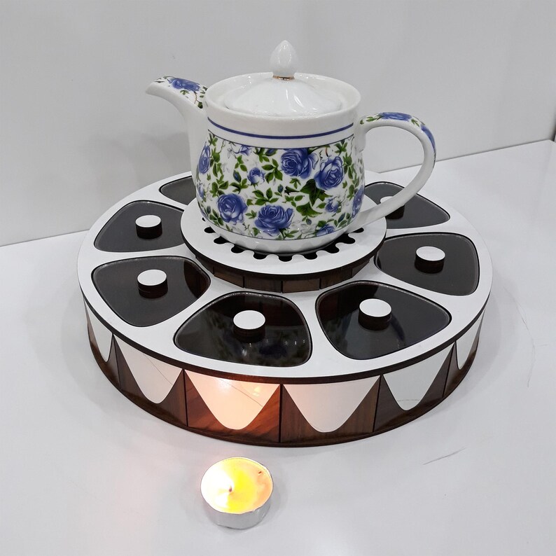 K&ouml;nnte beinhalten: Eine wei&szlig;e Teekanne aus Keramik mit Blumenmuster steht auf einem Holztablett mit sechs F&auml;chern. Das Tablett hat ein wei&szlig;-braunes Design. Im Vordergrund befindet sich eine kleine Kerze.