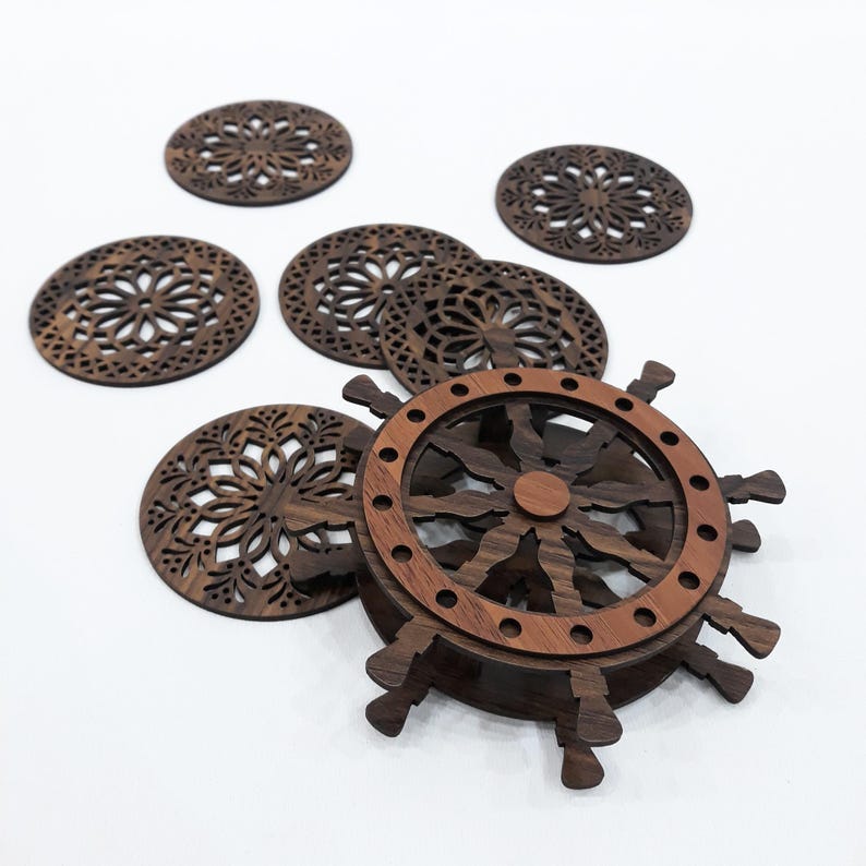 May include: A set of dark brown wooden coasters featuring intricate laser-cut floral patterns, accompanied by a coaster holder designed to resemble a ship's steering wheel with a lighter brown outer ring and dark brown spokes and base.