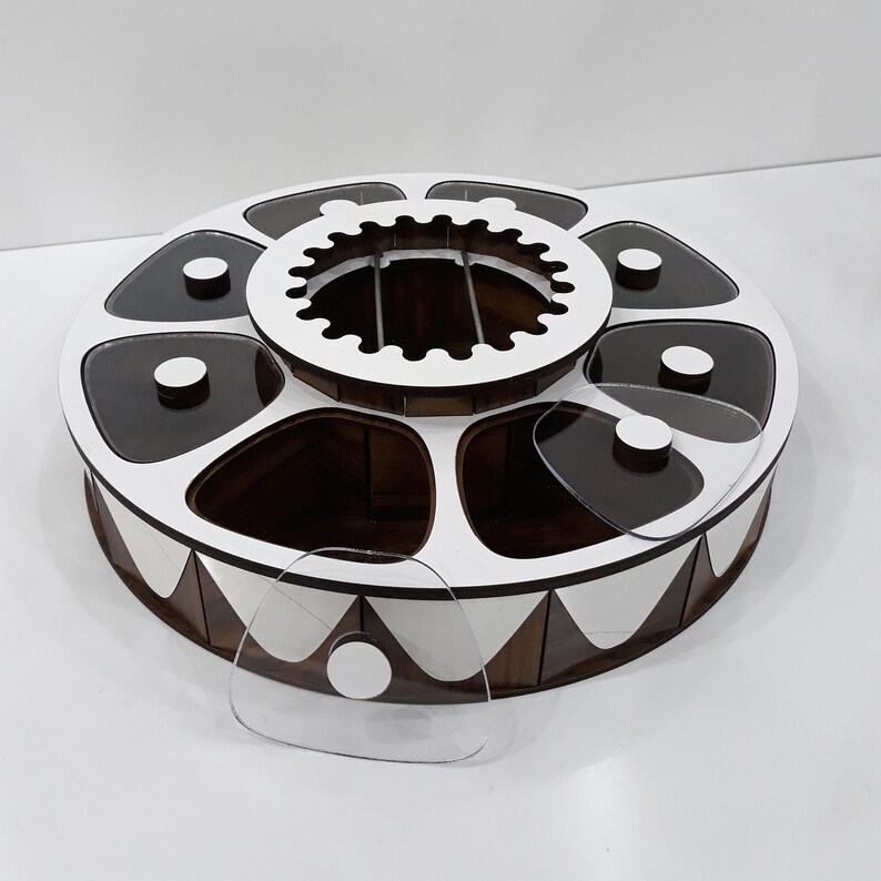 K&ouml;nnte beinhalten: Ein rundes Holztablett mit sechs einzelnen F&auml;chern, jedes mit einem durchsichtigen Deckel. Das Tablett hat ein wei&szlig;-braunes geometrisches Design und ein wei&szlig;es Mittelst&uuml;ck mit einem dekorativen Zahnrad-Design.