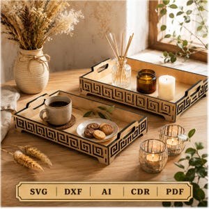 May include: Two rectangular wooden trays featuring a black laser-cut Greek key pattern along the sides and integrated handles. The trays hold a ceramic mug with a beverage, a small plate of cookies, a glass candle holder, and a reed diffuser.
