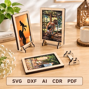 May include: Three light-colored wooden picture frames with dark edges, two standing upright and one lying flat, each displaying a different photograph. A separate wooden stand is placed nearby. The text SVG DXF AI CDR PDF is visible.