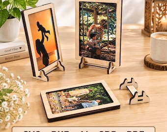 May include: Three light-colored wooden picture frames with dark edges, two standing upright and one lying flat, each displaying a different photograph. A separate wooden stand is placed nearby. The text SVG DXF AI CDR PDF is visible.