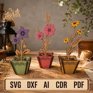 May include: Three laser-cut wooden flower decorations in square pots. The left pot is light green with a purple flower, the center is pink with a pink flower, and the right is yellow with a yellow flower. Text at the bottom reads: SVG DXF AI CDR PDF.
