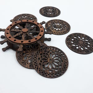 May include: A set of dark brown wooden coasters with intricate laser-cut floral patterns, accompanied by a circular storage holder shaped like a ship's wheel. The holder features a lighter brown rim with small circular cutouts along the edge.
