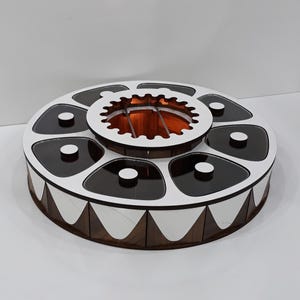 K&ouml;nnte beinhalten: Eine runde Holzkiste mit einem wei&szlig;en Deckel und einem zentralen Fach. Die Kiste hat sechs kleinere F&auml;cher am Rand, jedes mit einem schwarzen Deckel. Die Kiste hat ein wei&szlig;es und braunes geometrisches Muster am unteren Rand.