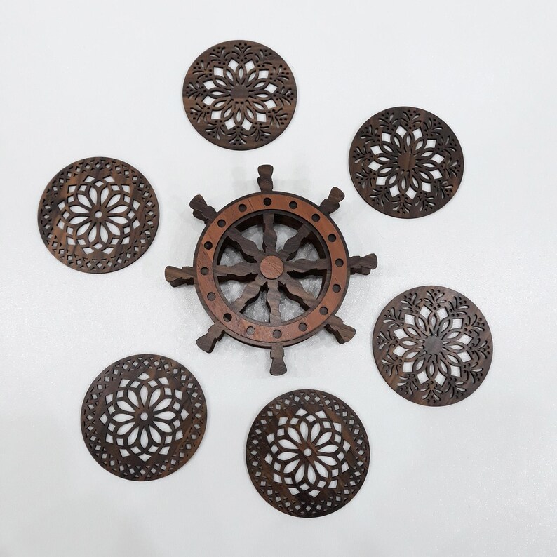 May include: A set of five circular dark brown wooden coasters featuring intricate laser-cut floral patterns, arranged around a central dark brown wooden holder shaped like a ship's steering wheel with eight spokes and a circular base.