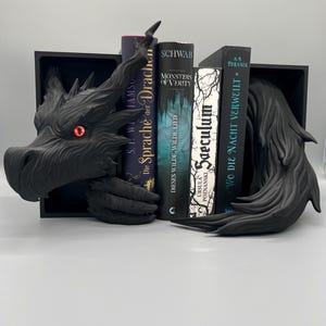 Könnte beinhalten: Ein schwarzer Drachen-Buchstütze mit roten Augen, die mehrere Bücher hält. Die Buchstütze besteht aus dunklem Material und weist komplizierte Details auf. Die Bücher haben verschiedene Titel und Farben, was einen Kontrast erzeugt.
