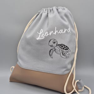Könnte beinhalten: Ein grauer Kordelzugrucksack mit braunem Boden. Der Rucksack hat den Namen "Leonhard" in weißer Schrift und eine schwarz-weiße Cartoon-Schildkröten-Illustration. Der Kordelzug ist cremefarben.