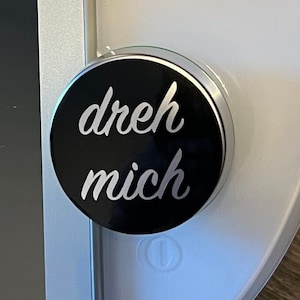 Könnte beinhalten: Ein runder schwarzer Knopf mit silbernem Rand. Der Knopf hat einen weißen Text, der "dreh mich" lautet, was im Deutschen "drehe mich" bedeutet.