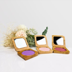 Könnte beinhalten: Drei Holz-Kompaktpuder mit Spiegeln und verschiedenen Rouge-Farbtönen. Die Kompaktpuder sind aus hellbraunem Holz und haben eine quadratische Form. Die Rouge-Farbtöne sind rosa, lila und hellrosa.