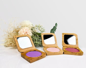 May include: Three wooden compacts with mirrors and different coloured blush shades. The compacts are made of light brown wood and have a square shape. The blush shades are pink, purple, and a light pink.