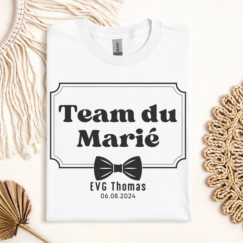 K&ouml;nnte beinhalten: Wei&szlig;es T-Shirt mit einer schwarz-wei&szlig;en Grafik, auf der "Team du Mari&eacute;" mit einer Fliege und dem Text "EVG Thomas 06.08.2024" steht.