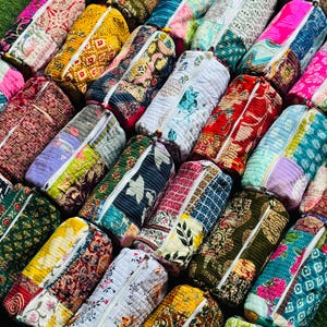 Op de afbeelding: Een verzameling cilindervormige, gewatteerde stoffen etuis met diverse kleurrijke patchworkpatronen, bloemenprints en geometrische ontwerpen. Elk etui is voorzien van een witte ritssluiting en heeft over het hele oppervlak gedetailleerd stikwerk.