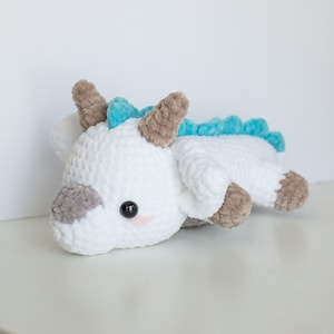 May include: A white crocheted plush toy in the shape of a cow, with gray and brown accents. The toy has a light blue spine and small horns. The cow is lying down on a white surface.
