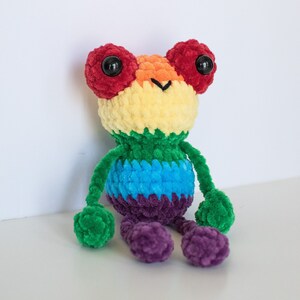 May include: A crocheted rainbow-coloured plush toy with large red eyes and a smiling face. The toy is sitting on a white surface.