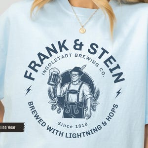 May include: Light blue t-shirt with a navy blue graphic. The graphic features the text "FRANK & STEIN INGOLDSTADT BREWING CO." around an illustration of a man holding a beer stein. The shirt also includes the text "BREWED WITH LIGHTNING & HOPS Since 1818."