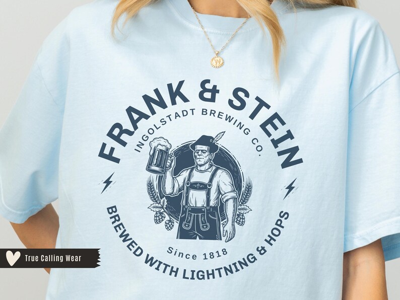 May include: Light blue t-shirt with a navy blue graphic. The graphic features the text "FRANK & STEIN INGOLDSTADT BREWING CO." around an illustration of a man holding a beer stein. The shirt also includes the text "BREWED WITH LIGHTNING & HOPS Since 1818."