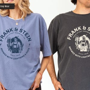 May include: Two t-shirts, one light blue and one charcoal gray, each featuring a circular graphic. The graphic includes the text "FRANK & STEIN INGOLSTADT BREWING CO." and an illustration of a man holding a beer stein. The text "BREWED WITH LIGHTNING & HOPS" is also present.