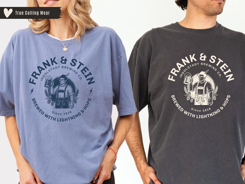 May include: Two t-shirts, one light blue and one charcoal gray, each featuring a circular graphic. The graphic includes the text "FRANK & STEIN INGOLSTADT BREWING CO." and an illustration of a man holding a beer stein. The text "BREWED WITH LIGHTNING & HOPS" is also present.
