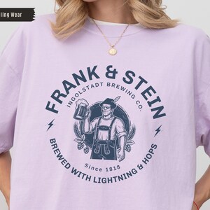 May include: A light purple t-shirt featuring a circular graphic for "Frank & Stein Ingolstadt Brewing Co." The design includes a man holding a beer stein, lightning bolts, and the text "Brewed with Lightning & Hops Since 1818."